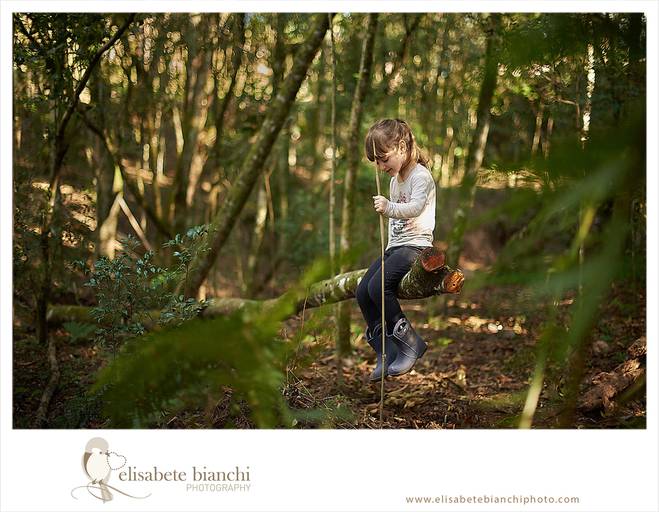 Criança com galocha e vareta, sentada em um galho de árvore, no meio da mata, em ensaio infantil externo, entre Caxias do Sul e Farroupilha.