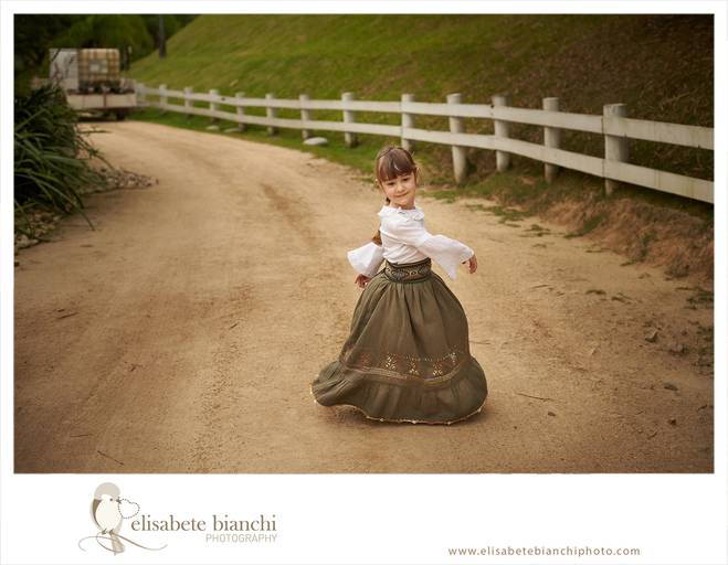 Criança vestida de prenda, ao lado de uma cerca branca de madeira, rodando saia típica do Rio Grande do Sul, em uma estrada de chão batido, em ensaio infantil externo, em Caxias do Sul.