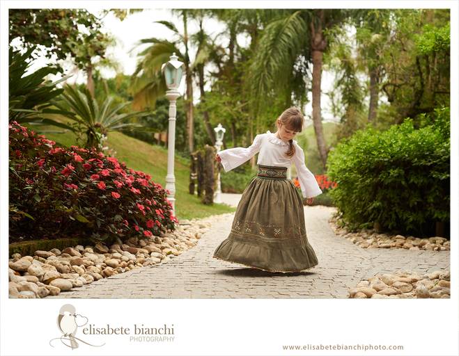 Criança vestida de prenda, rodando saia típica do Rio Grande do Sul, em meio a um jardim, em ensaio infantil externo, em Caxias do Sul.