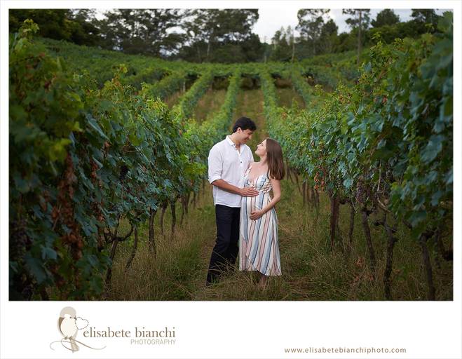 Casal gestante olhando-se nos olhos e acarinhando a barriga. Eles estão em meio a um parreiral. A fotografia faz parte do ensaio externo da gestação, realizado na Casa Tonet, em Caxias do Sul.