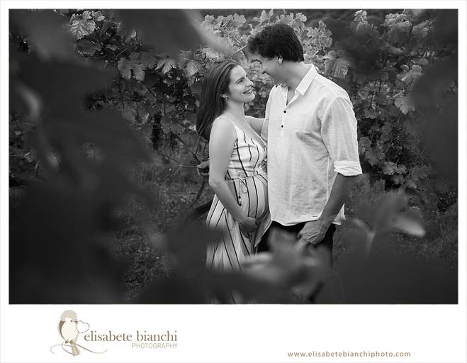 Casal gestante olhando-se nos olhos abraçados. Eles estão emoldurados com as folhas da videira. A fotografia faz parte do ensaio externo da gestação, realizado na Casa Tonet, em Caxias do Sul.