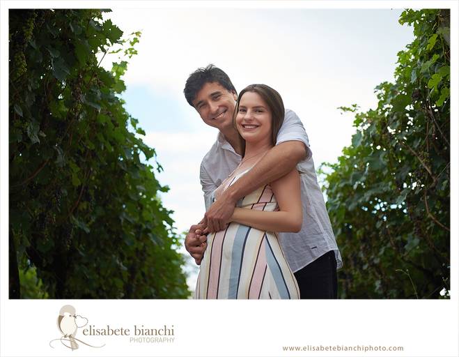 Casal gestante olhando para a câmera, abraçados. Há videiras nas duas laterais e  ao fundo do casal , o céu, os deixando em destaque. A fotografia faz parte do ensaio externo da gestação, realizado na Casa Tonet, em Caxias do Sul.