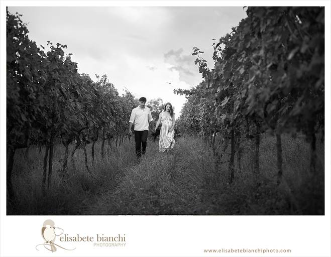 Casal gestante caminha em direção à câmera, entre videiras. A fotografia faz parte do ensaio externo da gestação, realizado na Casa Tonet, em Caxias do Sul. 