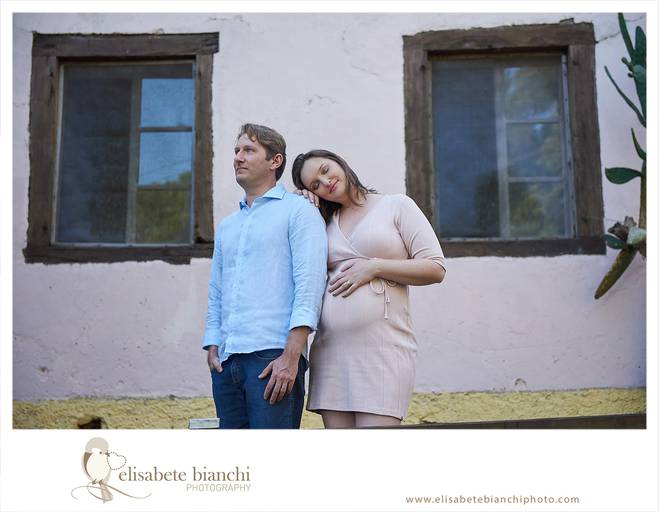 Pai e mãe gestante, lado a lado, refletindo. O pai olha o horizonte e a mãe descansa a cabeça em seu ombro. Eles estão em frente à fachada da Casa Bonnet, no interior de Caxias do Sul, em ensaio de fotos da gestação.