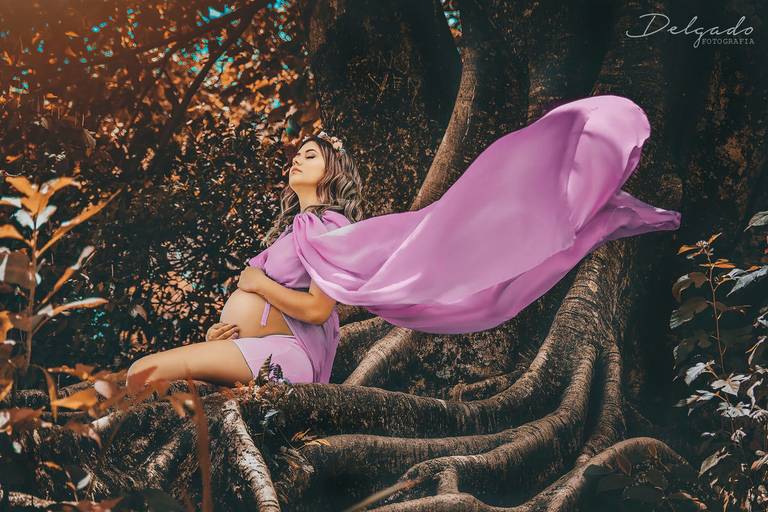 gestante sentada nas raízes de uma árvore com a calda de seu vestido voando ao vento