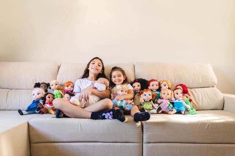 Meninas sentadas no sofá com bebê no colo e pelúcias das princesas 