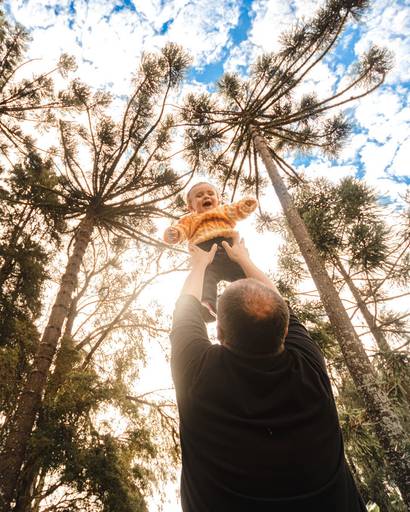 pai brincando levantando filho para  no parque com árvores no fundo