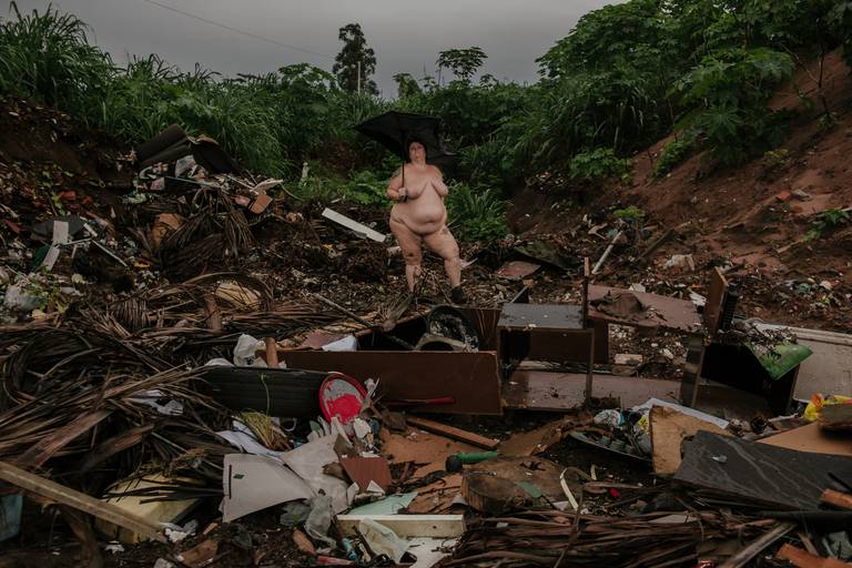foto na horizontal com uma mulher branca, gorda e nua segurando um guarda-chuva quebrado. ela está no meio do lixo e de restos de móveis no primeiro plano