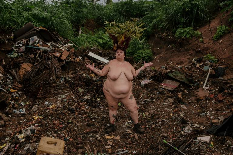 foto na horizontal com uma mulher branca, gorda e nua ao centro, usando uma coroa de flores e galhos em tons de vermelho, verde e marrom. ela está com os braços semi levantados e expressão de questionamento. é possível ver restos de lixo e terra ao redor