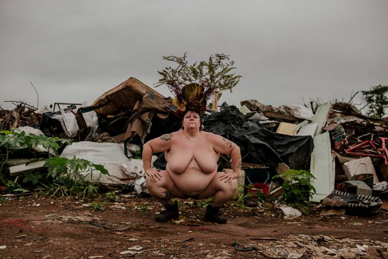 foto na horizontal com uma mulher branca, gorda e nua, usando uma coroa de flores e galhos em tons de vermelho, marrom e verde. ela está agachada com as mãos apoiadas nos joelhos e é possível ver uma montanha de lixo e restos de móveis ao fundo
