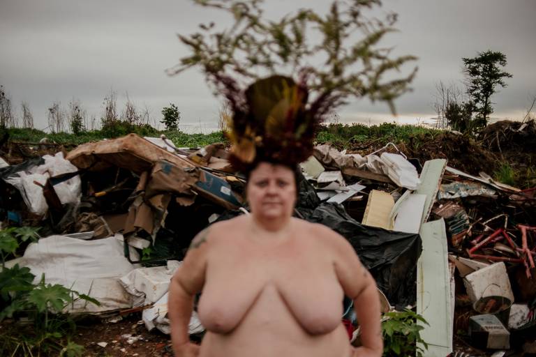 foto na horizontal com uma mulher branca, gorda e nua ao centro usando uma coroa de flores e galhos em tons de marrom, verde e vermelho. ela está desfocada, trazendo a atenção para a pilha de lixos ao fundo dela