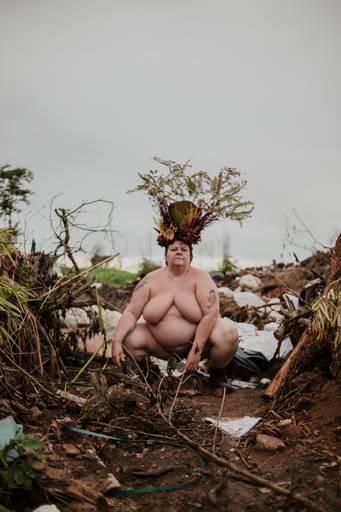 foto na horizontal com uma mulher branca, gorda e nua, usando uma coroa de flores e galhos em tons de vermelho, marrom e verde. ela está agachada com um dos braços mais a frente e é possível ver uma montanha de lixo e terra ao fundo