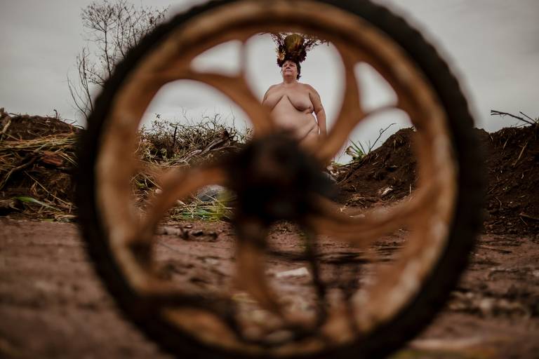 foto na horizontal com uma roda em primeiro plano desfocada e uma mulher gorda, branca e nua usando uma coroa de flores vermelha, marrom e verde na parte superior da imagem, entre os raios da roda. ao fundo há um monte de terra