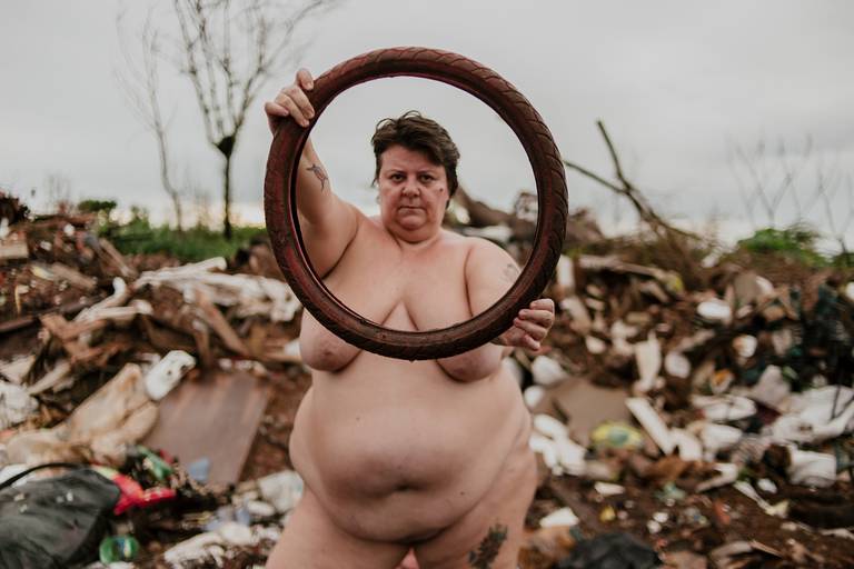 foto na horizontal com uma mulher branca, gorda e nua segurando um pneu de bicicleta com as duas mãos e seu rosto está dentro do círculo do pneu. é possível ver uma montanha de lixo desfocada ao fundo