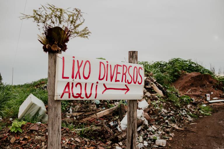 foto na horizontal com uma placa branca apoiada em dois pilares de madeira com a frase lixo diversos aqui, uma coroa de flores e galhos apoiado no pilar de madeira da esquerda e ao fundo uma montanha de terra e lixo
