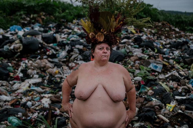 foto na horizontal com uma mulher gorda, branca e nua ao centro, visto da cintura para cima, usando uma coroa de flores e galhos nas cores vermelho, marrom e verde e ao fundo uma montanha de lixo