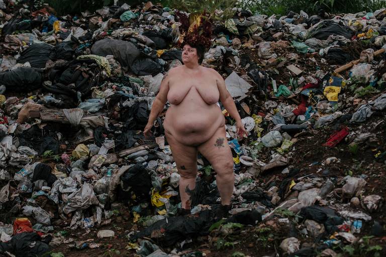 foto na horizontal com uma mulher gorda, branca e nua ao centro, de corpo inteiro e com os braços semiabertos, usando uma coroa de flores e galhos nas cores vermelho, marrom e verde e ao fundo uma montanha de lixo