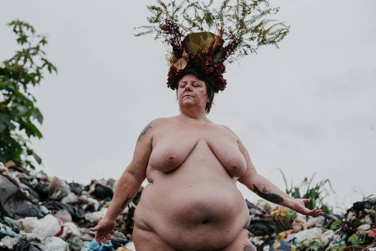 foto na horizontal com uma mulher gorda, branca e nua ao centro, de olhos fechados e a mão direita levemente erguida e vista da barriga pra cima, usando uma coroa de flores e galhos nas cores vermelho, marrom e verde e ao fundo uma montanha de lixo