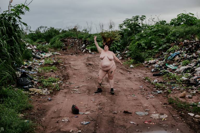 foto na horizontal de uma mulher branca, gorda e nua, com botas marrons nos pés e uma coroa de flores e galhos vermelhos, verdes e marrons na cabeça, com o braço esquerdo levantado, em meio a uma estrada de terra com montanhas de lixo nas laterais