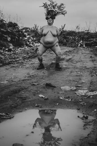 foto preto e branco na vertical com uma mulher branca, gorda e nua, em posição semiagachada com os dois braços levantados, no meio de uma estrada de terra com lixos espalhados. é possível ver o reflexo da modelo na poça de lama na parte inferior da imagem