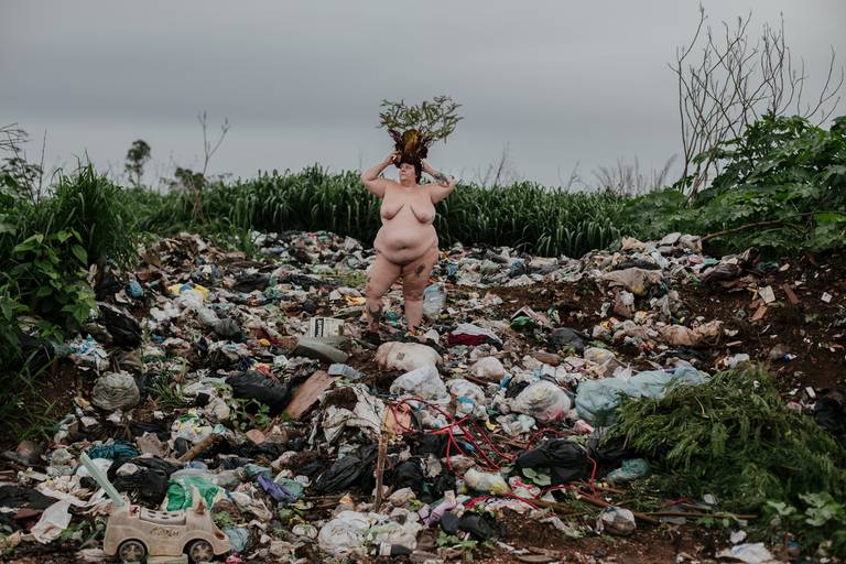 foto na horizontal com uma mulher branca, gorda e nua segurando com as duas mãos uma coroa de flores e galhos na cabeça em tons de vermelho, verde e marrom. ela está centralizada no meio da imagem em meio à uma montanha de lixo