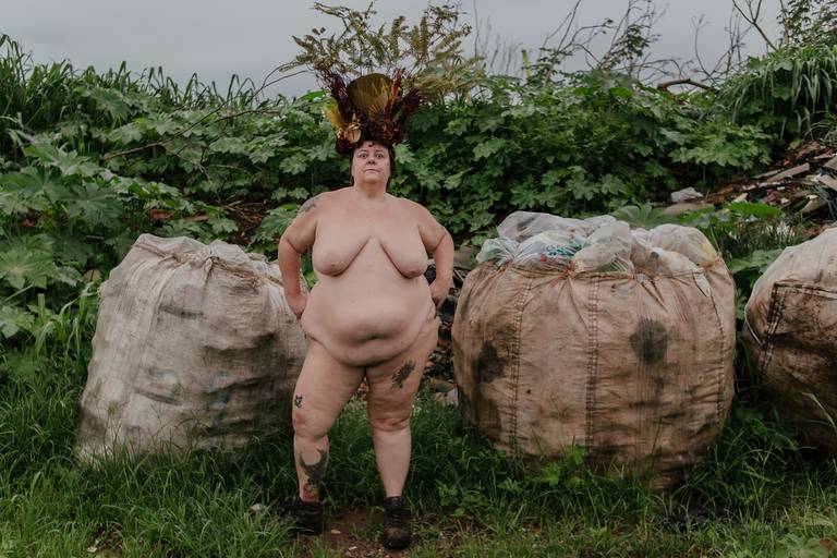 foto na horizontal com uma mulher branca, gorda e nua ao centro, usando uma coroa de flores e galhos em tons de vermelho, verde e marrom e com dois sacos grandes de lixo de cada lado e ao fundo um mato verde