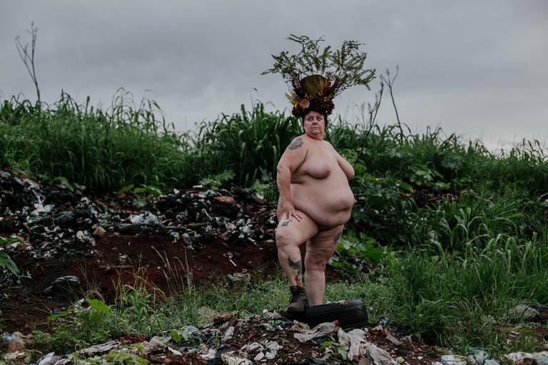 foto na horizontal com uma mulher gorda, branca e nua no centro-direito, com o pé esquerdo apoiado em um pneu e a mão apoiada na coxa, usando uma coroa de flores e galhos nas cores vermelho, marrom e verde e ao fundo uma montanha de lixo