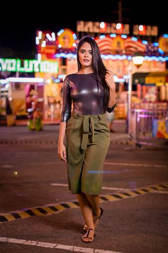 fotografia de moda colecao outono inverno em parque de diversoes modelo morena cabelos lontos calça caqui e blusa preta brilhante com luzes coloridas em night shot fotografia noturna ao fundo